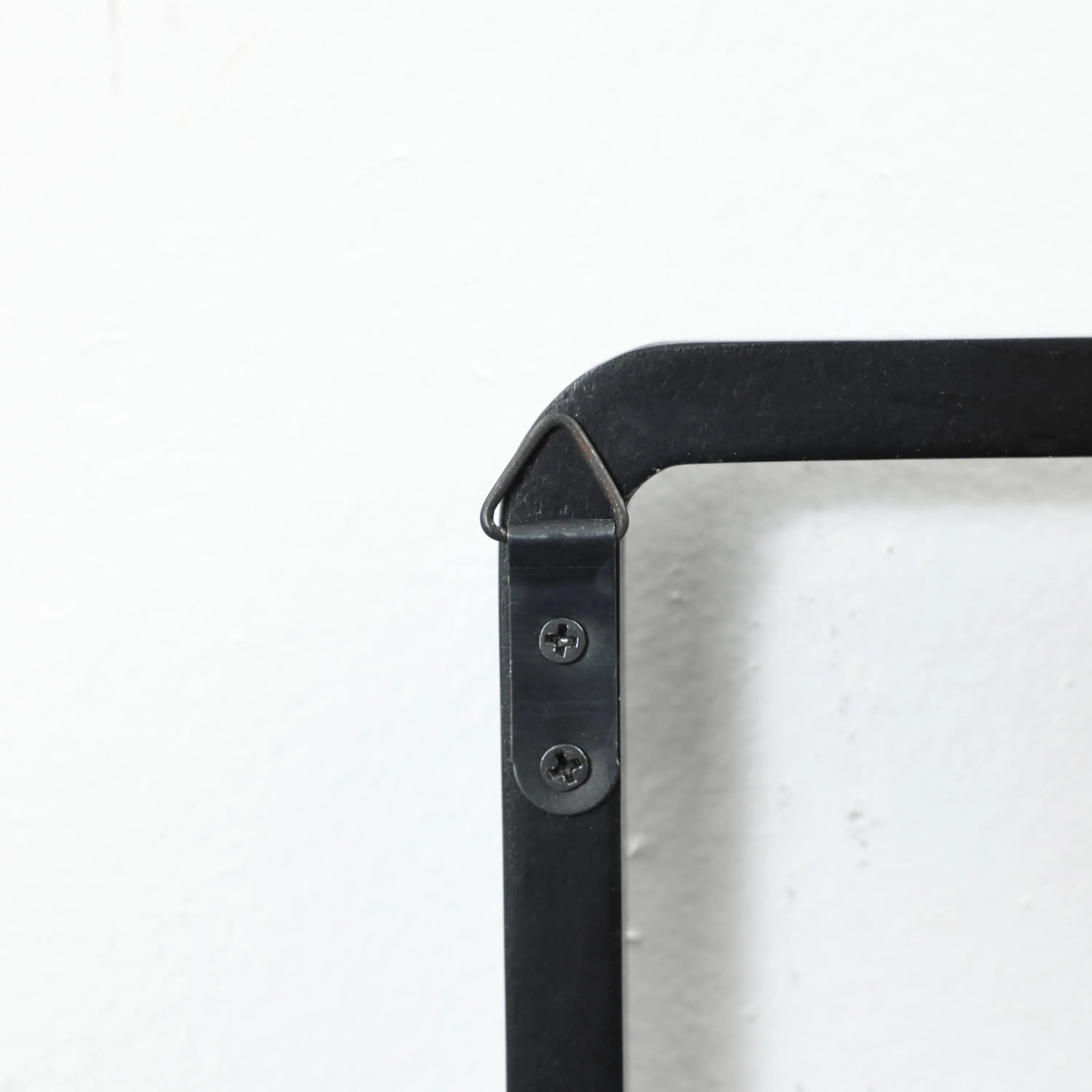 Close-up of a black, triangular metal bracket with two screws on a white wall, designed as part of a wall-mounted coat rack with flip-down hooks, securely connecting two metal pieces at a right angle.