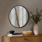 A round wall mirror with a wavy gold accent graces the space above a wooden sideboard, adding an artistic design touch. The sideboard elegantly displays books, a small vase with branches, and a decorative bowl, creating a modern decoration arrangement in the room.