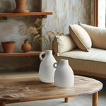 Two white ceramic and vintage vases with dried flowers grace a wooden coffee table in a cozy living room. The background features a beige couch and pottery on shelves, creating an inviting home decor ambiance.