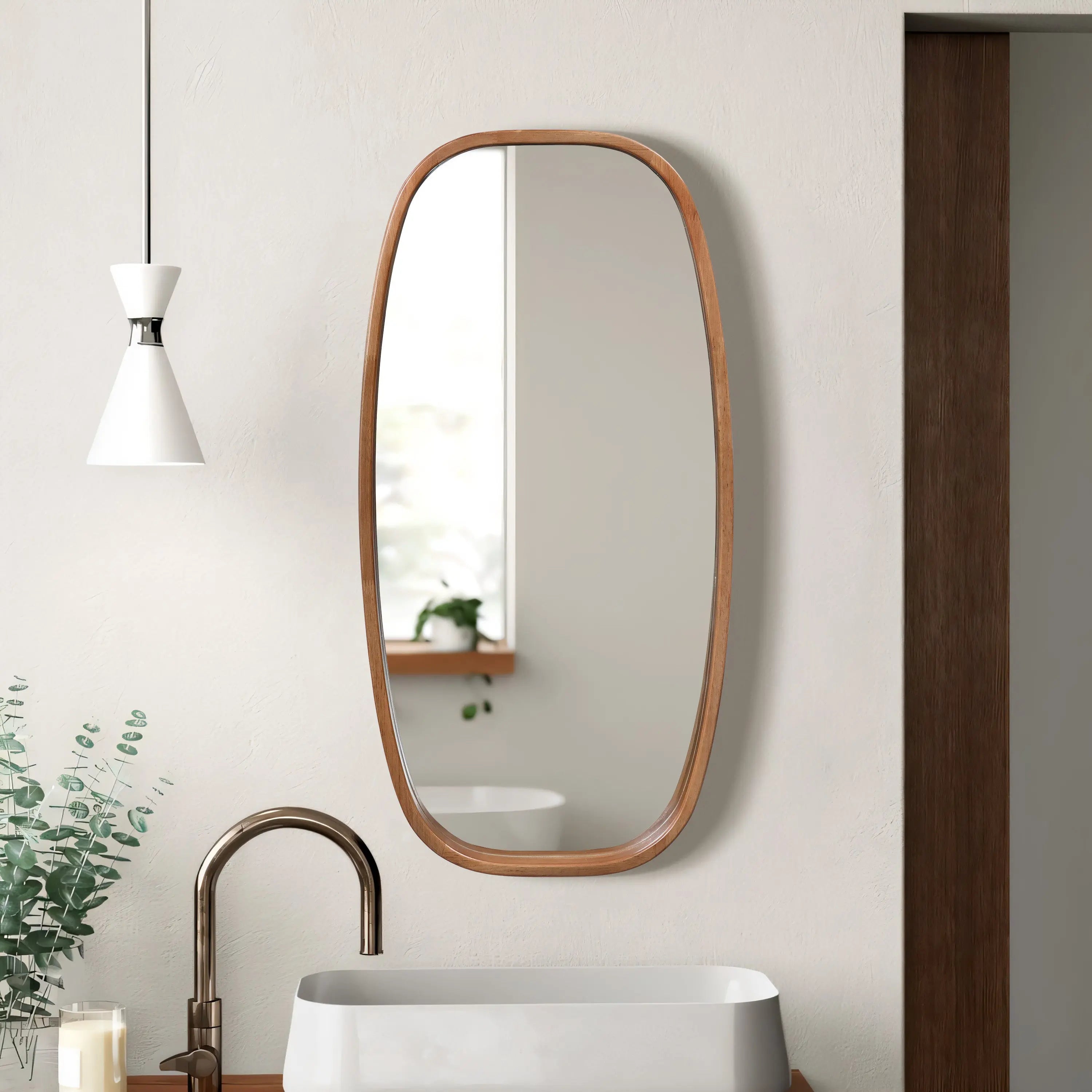 A modern wall mirror with an oval wooden frame hangs on a white wall above the sink, accompanied by a white pendant light and a planter brimming with greenery.