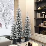 Three flocked artificial trees with realistic PVC branches in black pots are arranged in a modern living room with bookshelves and candles, while a snowy landscape is visible through the window.