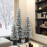 Three flocked artificial trees with realistic PVC branches in black pots are arranged in a modern living room with bookshelves and candles, while a snowy landscape is visible through the window.