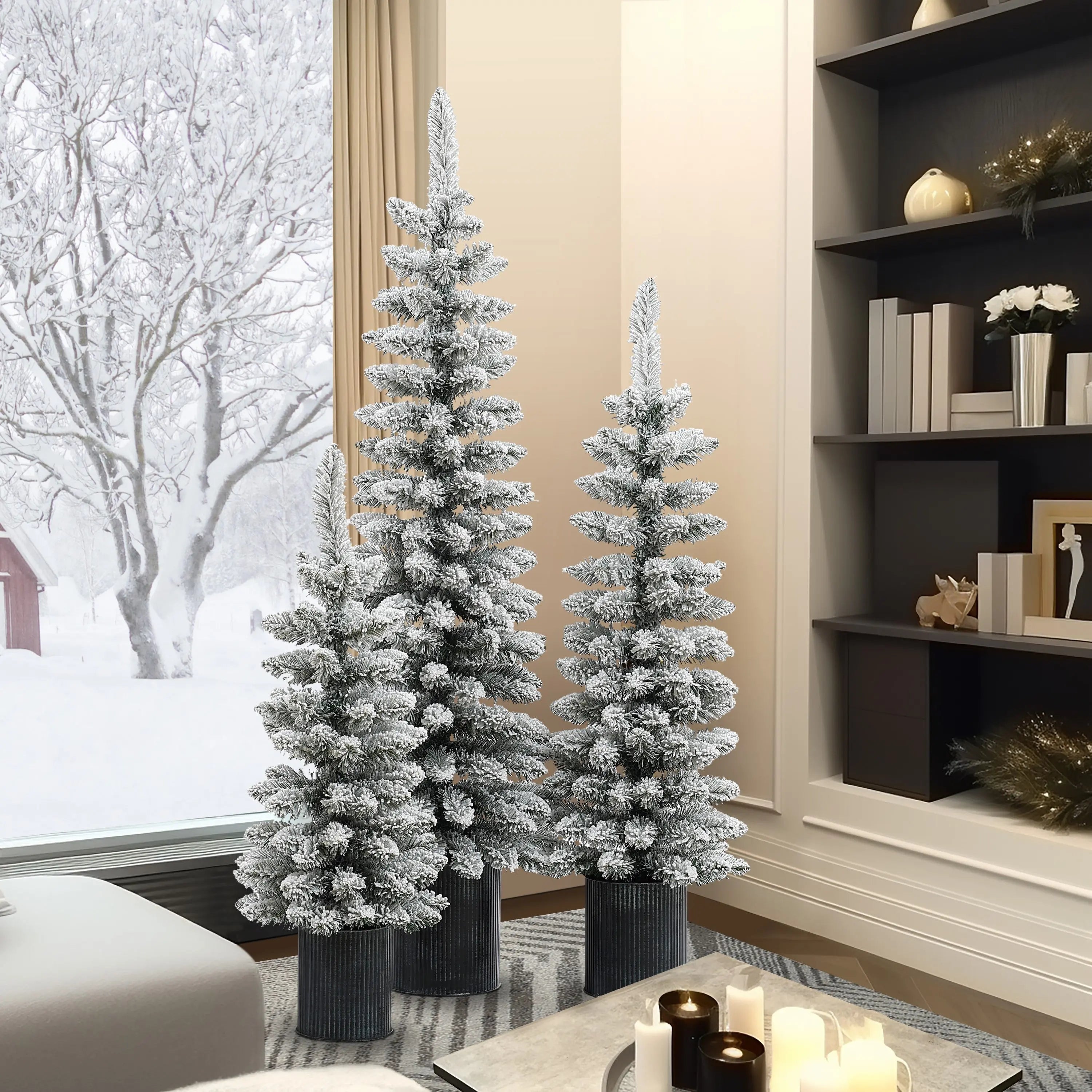 Three flocked artificial trees with realistic PVC branches in black pots are arranged in a modern living room with bookshelves and candles, while a snowy landscape is visible through the window.