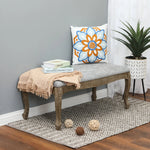 A wooden bench with linen upholstery and a gray cushion, topped with a floral pillow, sits on a patterned rug; a potted plant, books, and a beige throw add old-world charm to the decor.