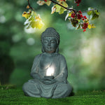 A stone Buddha statue sits cross-legged on grass, holding a small solar-powered light. Overhanging branches with leaves and red berries are above, making this serene scene a perfect garden decoration against a blurred green background.
