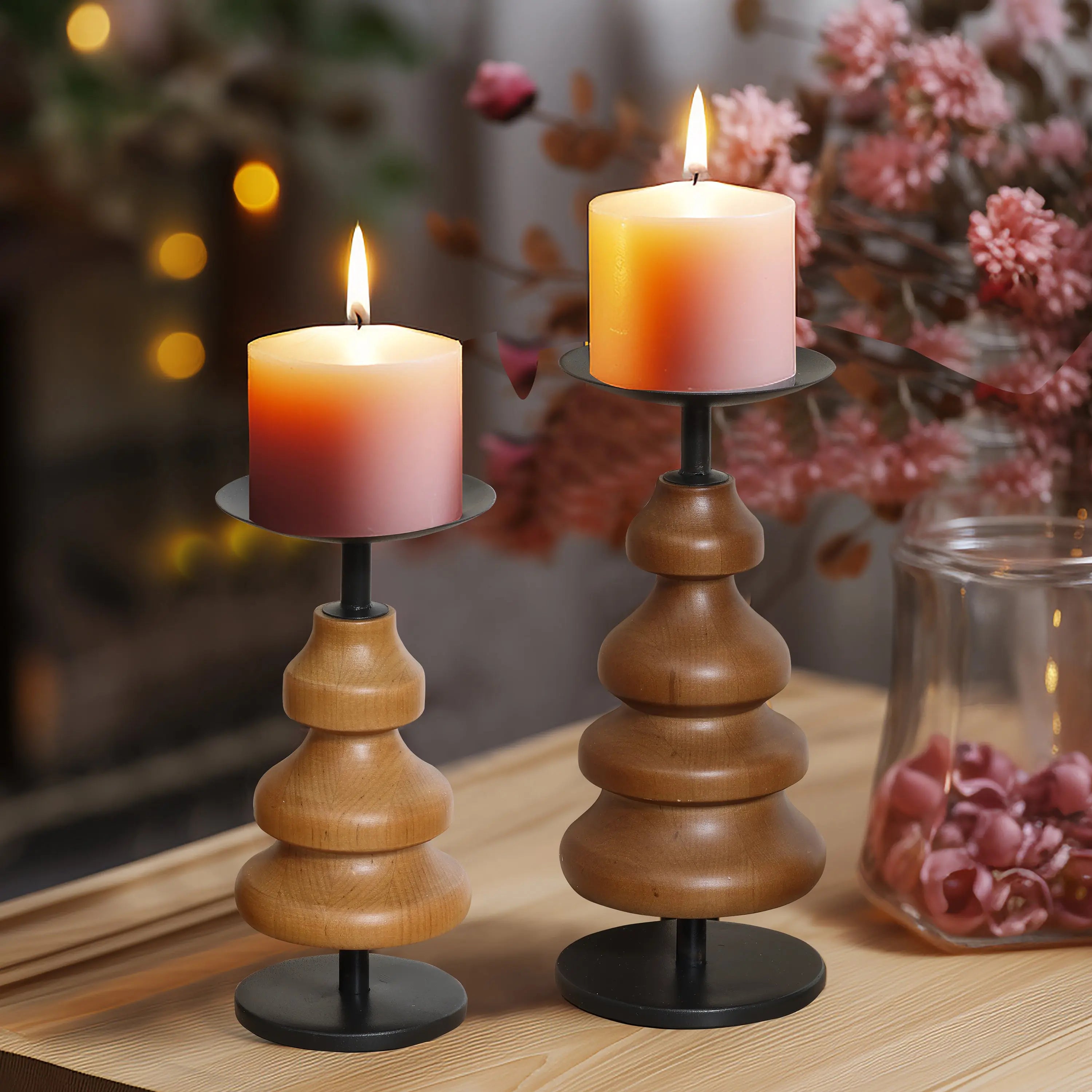 Two lit candles on birch wood holders sit elegantly on a table beside a jar of dried flowers, with blurred twinkling lights in the background, creating the perfect setting for cozy home decor.
