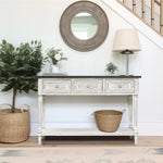 A farmhouse-inspired table with three drawers stands against the wall, topped with decor and a lamp; a round mirror hangs above, with woven baskets and a plant nearby.