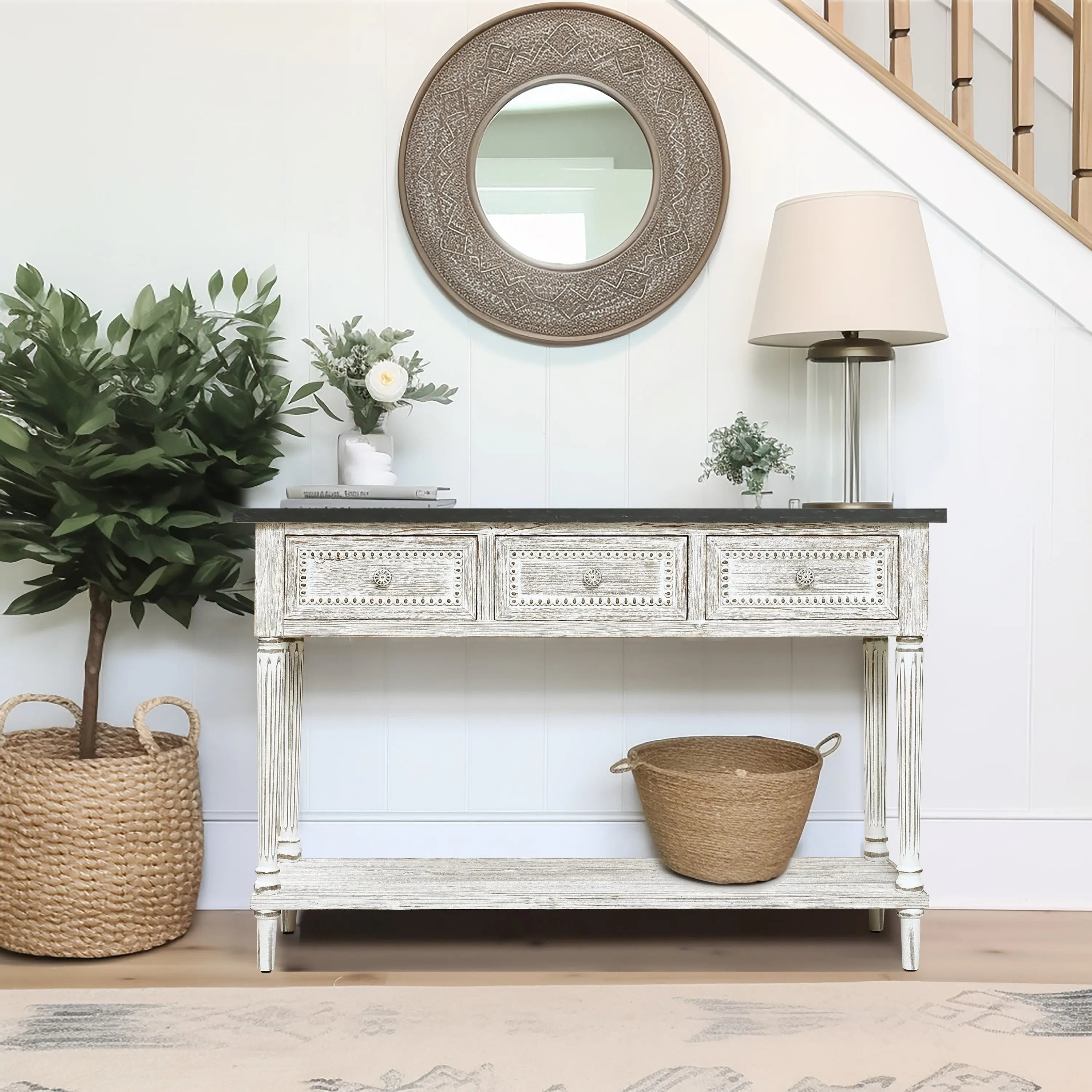 A farmhouse-inspired table with three drawers stands against the wall, topped with decor and a lamp; a round mirror hangs above, with woven baskets and a plant nearby.