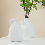 Two white speckled ceramic vases on a white table, one holding green leafy branches, bring a touch of vintage charm to your home decor against a beige background.