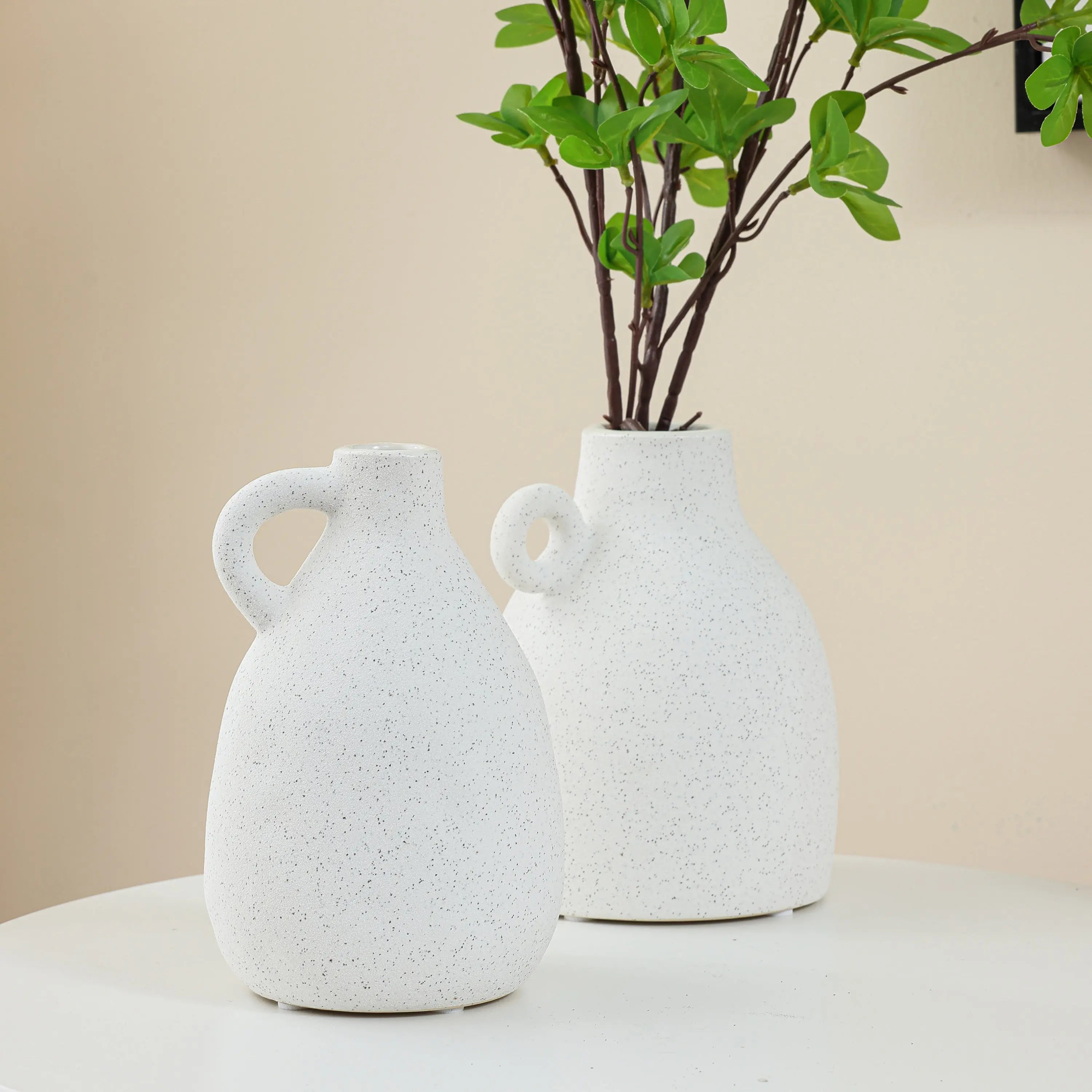 Two white speckled ceramic vases on a white table, one holding green leafy branches, bring a touch of vintage charm to your home decor against a beige background.