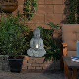 A Buddha statue holding a solar-powered light sits among green plants in a patio garden, next to an outdoor chair and a blue candle—a peaceful garden decoration.