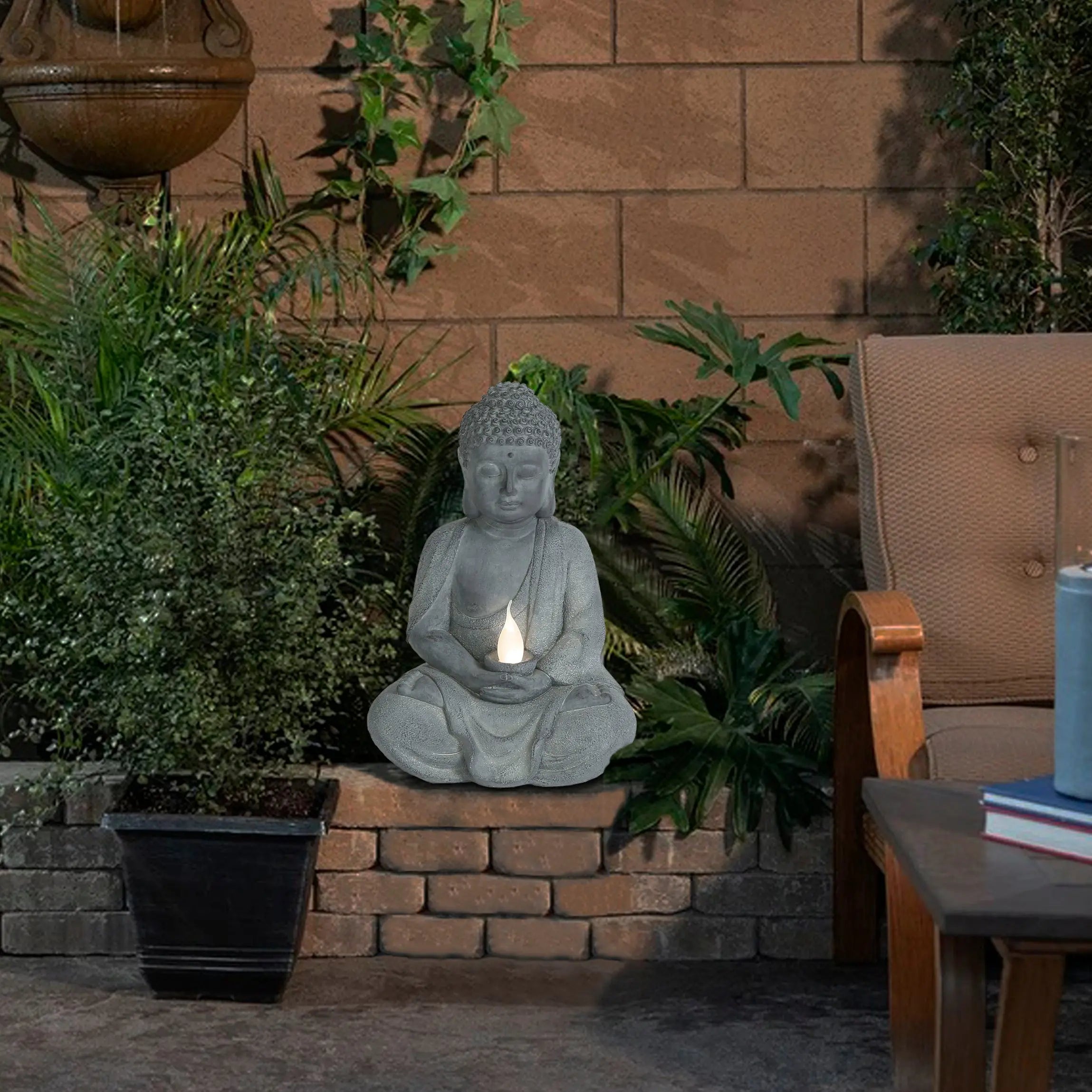 A Buddha statue holding a solar-powered light sits among green plants in a patio garden, next to an outdoor chair and a blue candle—a peaceful garden decoration.