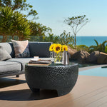 A black cement outdoor coffee table adorned with sunflowers, a silver coffee pot, and a cup. In the background, a gray sofa complements the scenic ocean view.