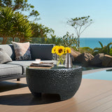 A black cement outdoor coffee table adorned with sunflowers, a silver coffee pot, and a cup. In the background, a gray sofa complements the scenic ocean view.