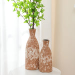 Two textured brown ceramic vases, exuding rustic charm and handcrafted earthenware appeal, stand on a white round table. The taller vase contains green leafy branches, adding a vibrant touch to the serene setting.