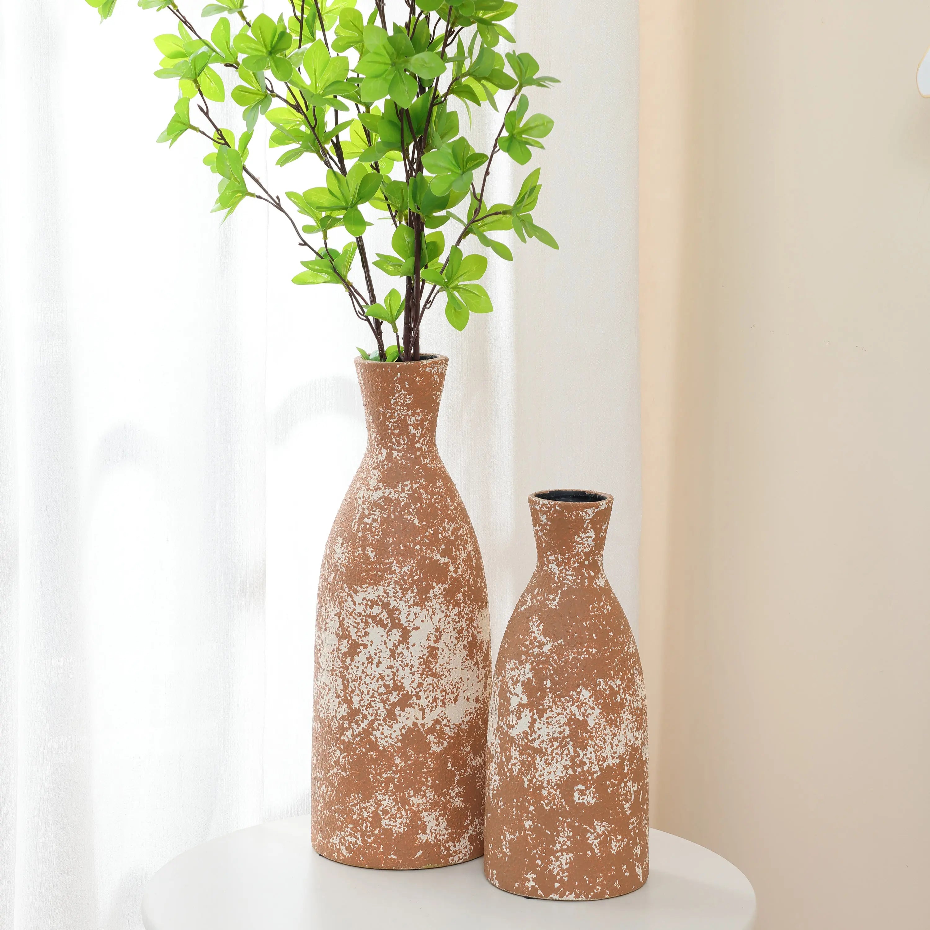 Two textured brown ceramic vases, exuding rustic charm and handcrafted earthenware appeal, stand on a white round table. The taller vase contains green leafy branches, adding a vibrant touch to the serene setting.