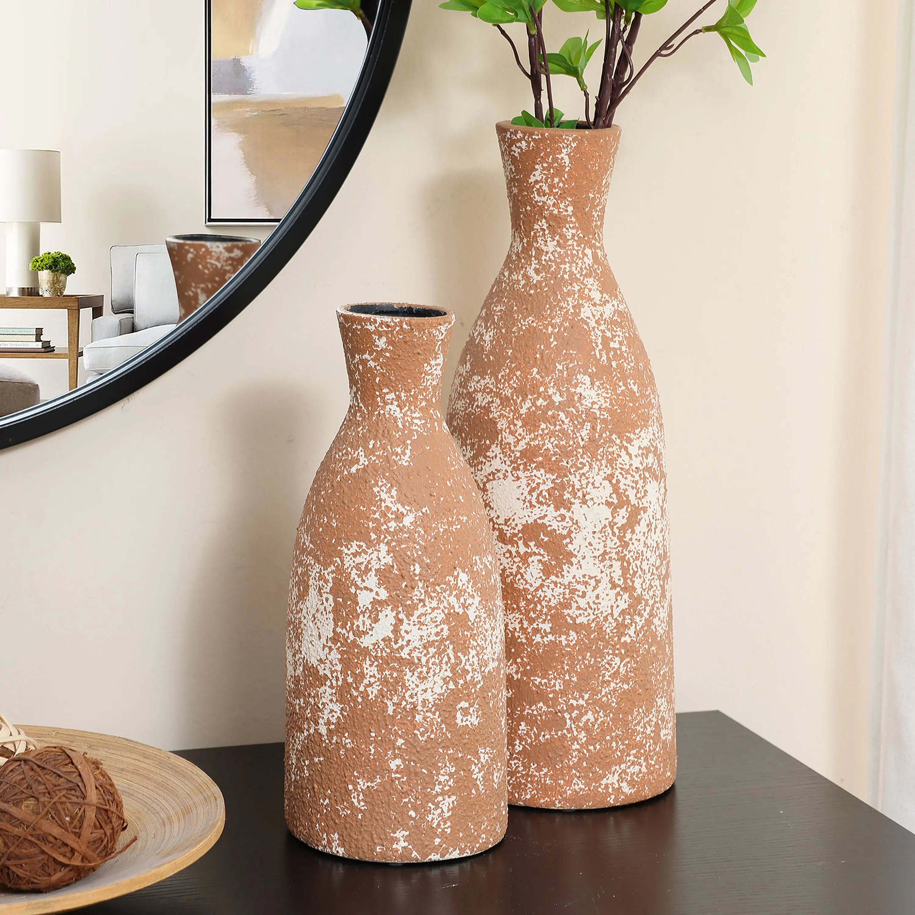 Two tall brown vases, exuding rustic charm with their white speckles, stand on a dark tabletop. One handcrafted earthenware vase cradles green branches. Nearby, a round mirror and a wooden bowl complete the aesthetic tableau.