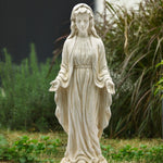 A durable magnesium oxide Mary statue in flowing robes stands outdoors with arms slightly extended, surrounded by grass and plants, making a serene religious icon for any garden.