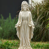 A durable magnesium oxide Mary statue in flowing robes stands outdoors with arms slightly extended, surrounded by grass and plants, making a serene religious icon for any garden.