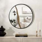 Round mirror with a black frame reflects a bedroom with pillows and a wall sconce. The left half of the mirror features an artistic design with a decorative golden crack. Two candles and books enhance the modern decoration on the console table.