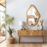 Modern living room with a natural wooden console table, a wavy-framed mirror, and a potted plant adding an organic touch. Versatile accent pieces complement the scene, while the reflection reveals a beautifully decorated bedroom.
