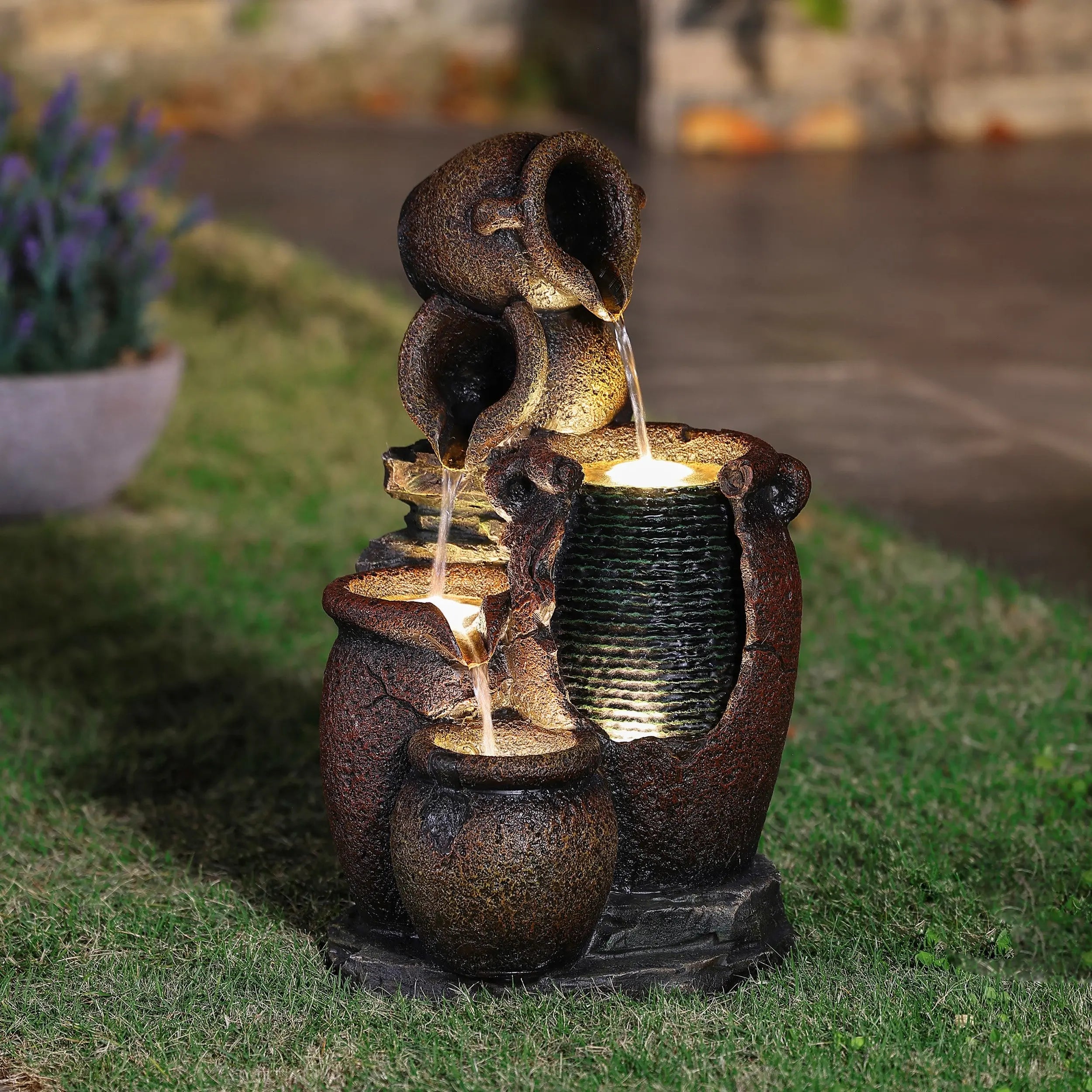 A rustic urn outdoor fountain featuring three jugs and an LED light in the illuminated basin, set on grass in a garden at dusk—an inviting piece of garden decor.