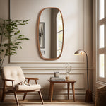 A cozy living room features a tall, modern wall mirror on the wall above a small wooden table. A beige armchair sits nearby, with a floor lamp and a leafy plant completing the decor.