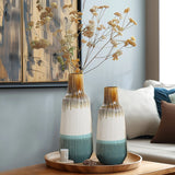Two unique ceramic vases with dried flowers sit on a wooden tray on a table, next to a cushioned sofa and abstract wall art.