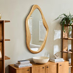 A natural, wood-framed mirror with an irregular shape adorns a beige wall, surrounded by wooden shelves, books, a bowl, and a potted plant. This organic addition serves as a versatile accent to any room.