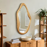 A natural, wood-framed mirror with an irregular shape adorns a beige wall, surrounded by wooden shelves, books, a bowl, and a potted plant. This organic addition serves as a versatile accent to any room.