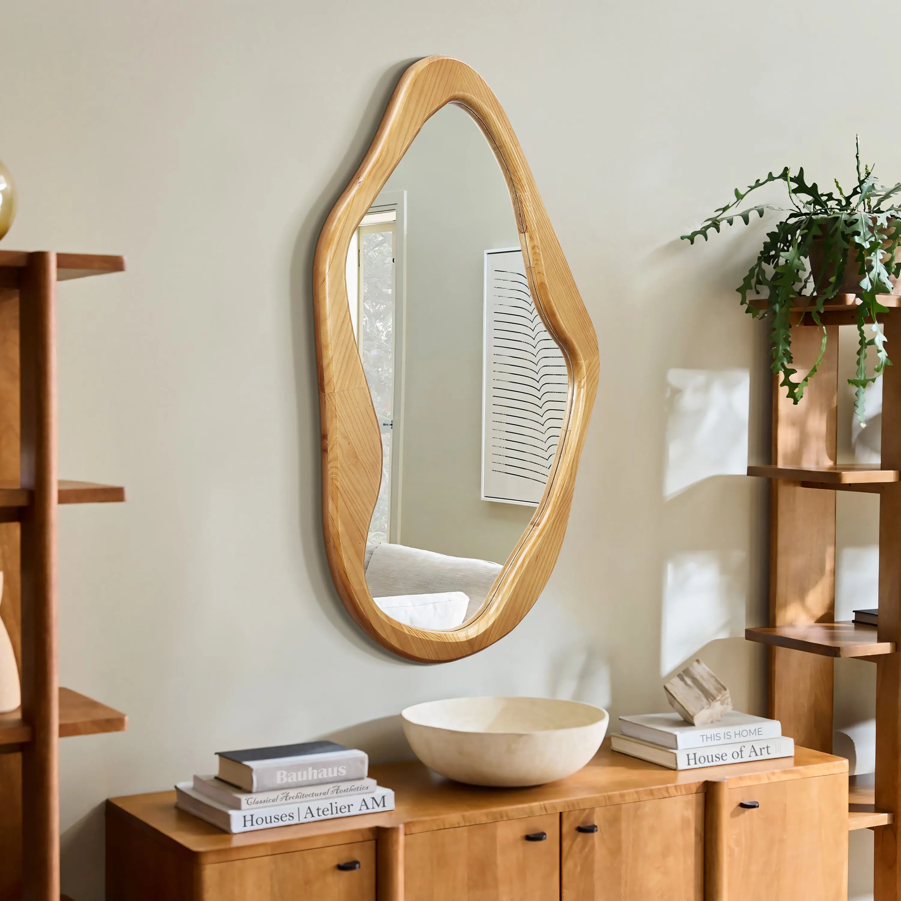 A natural, wood-framed mirror with an irregular shape adorns a beige wall, surrounded by wooden shelves, books, a bowl, and a potted plant. This organic addition serves as a versatile accent to any room.