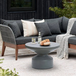 A modern outdoor seating area with a dark gray sofa and a contemporary round coffee table in a sleek gray finish, holding drinks and fruit, atop a light rug on a wooden deck with greenery in the background.