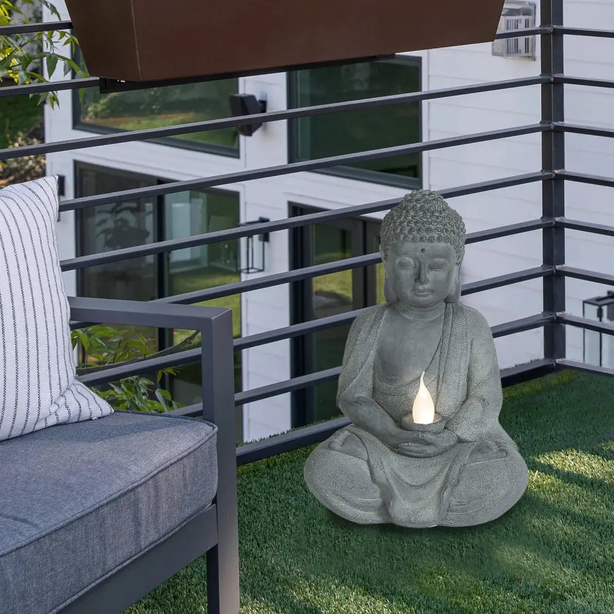 A Buddha statue holding a solar-powered light sits on artificial grass beside a metal railing and outdoor sofa, making a serene garden decoration for the balcony.