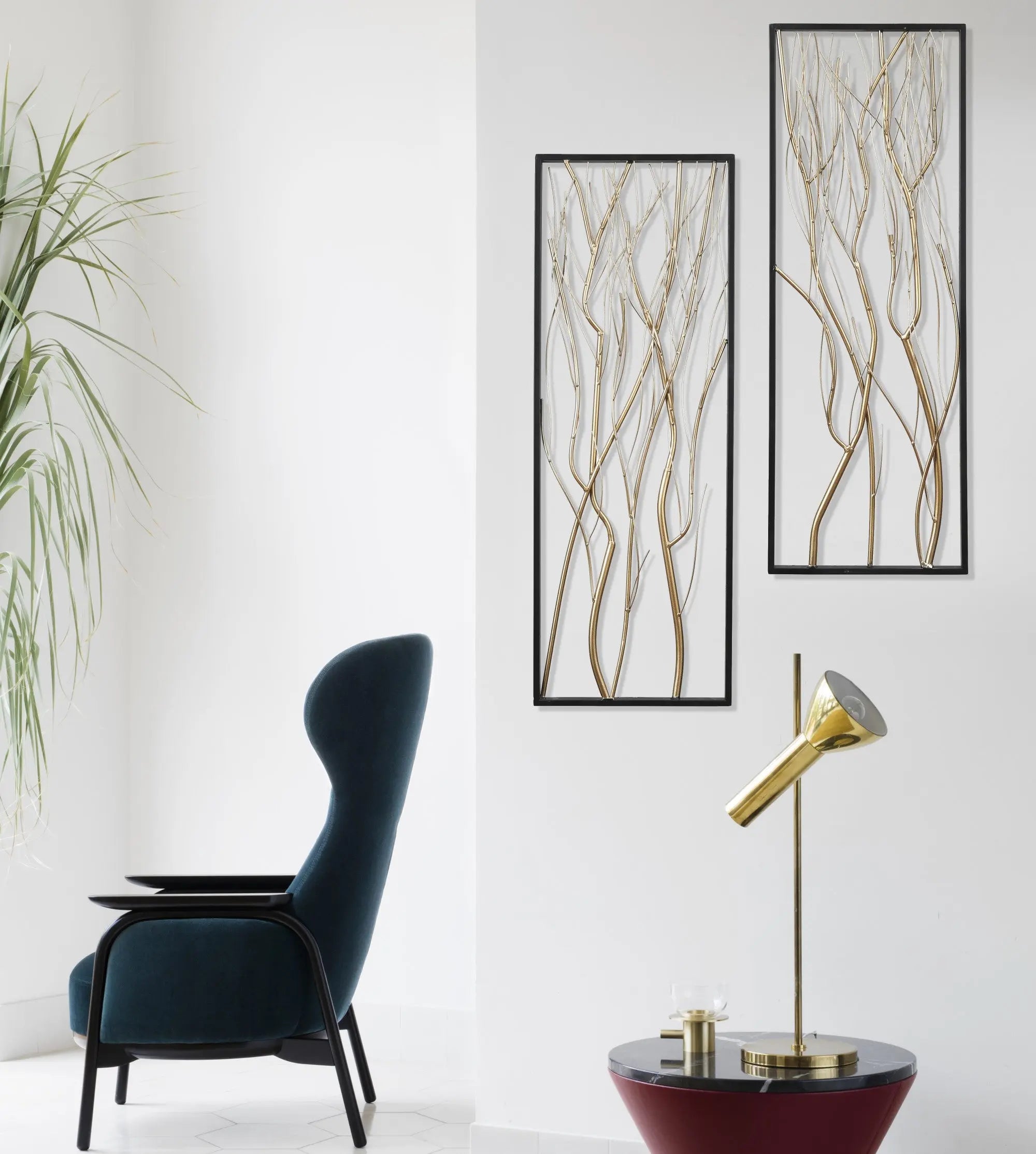 A modern living space featuring a dark blue armchair, a gold desk lamp on a round table, a potted plant, and striking modern wall art that serves as a focal point with its gold and silver branch designs.