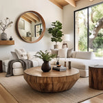Stylish living room with a round wooden coffee table boasting a natural wood finish, white sofa, large mirror, indoor plant, and a window view of trees. Neutral tones and modern decor create a cozy atmosphere.