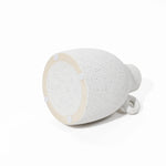 A speckled white ceramic vase featuring a vintage-inspired design lies on its side, showcasing a narrow neck and round base, as seen from the bottom.