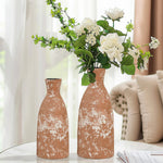 Two rustic brown vases, including a tall round ceramic vase, sit on a white table, one filled with white flowers and greenery. A beige sofa and sheer curtain complete the scene, adding rustic charm to the background.