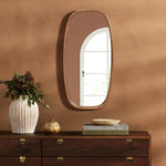 A handcrafted rubberwood mirror hangs on the brown wall above a dark wooden dresser adorned with books and vases. The windows reflection graces this contemporary style mirror, adding depth and elegance to the space.