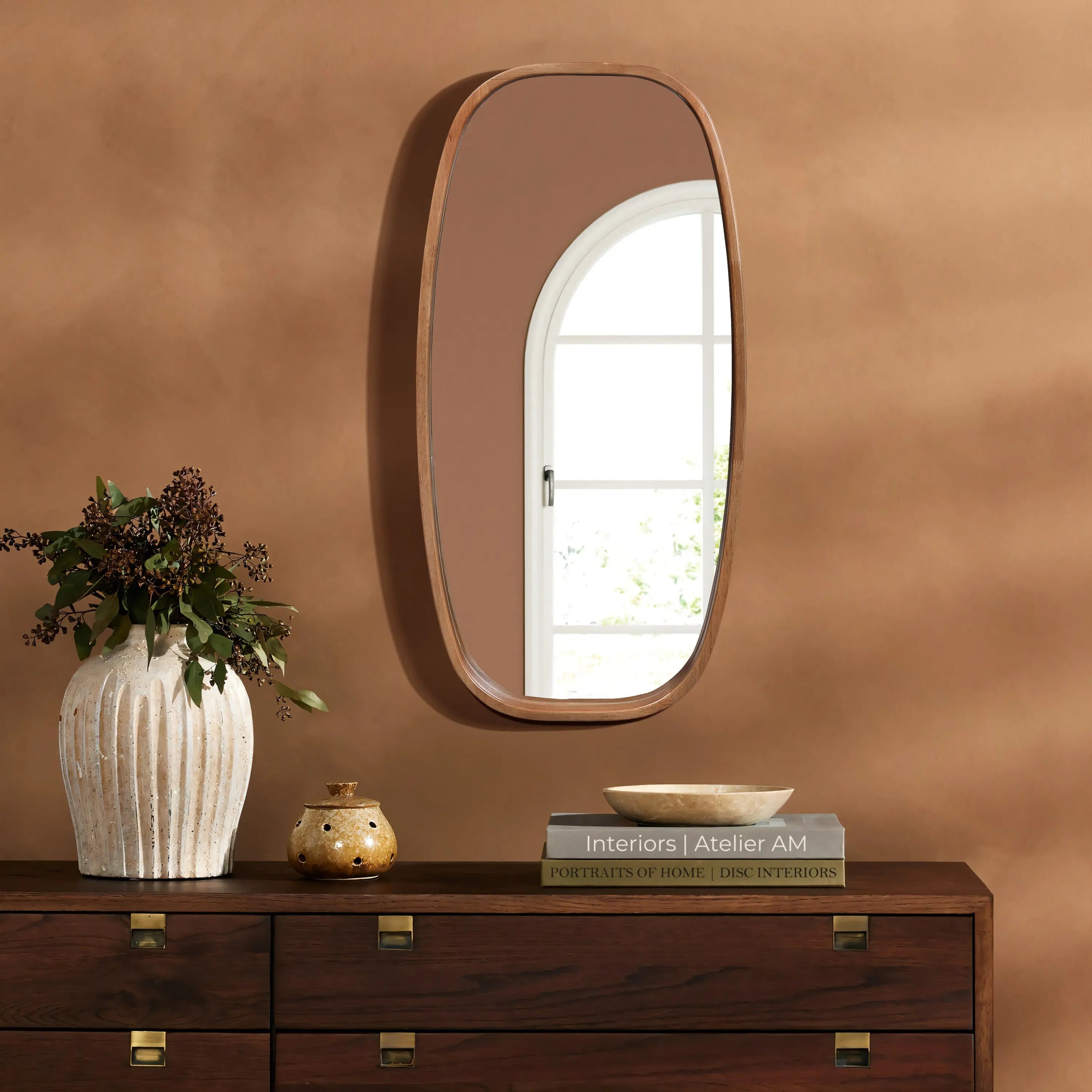 A handcrafted rubberwood mirror hangs on the brown wall above a dark wooden dresser adorned with books and vases. The windows reflection graces this contemporary style mirror, adding depth and elegance to the space.