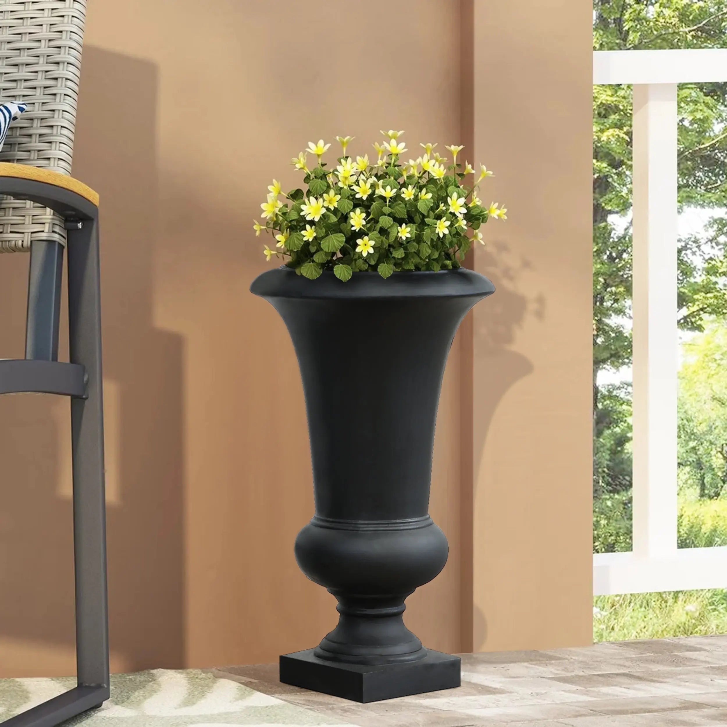 A weather-resistant black urn planter with yellow flowers sits on a porch beside a chair with a woven seat and backrest.