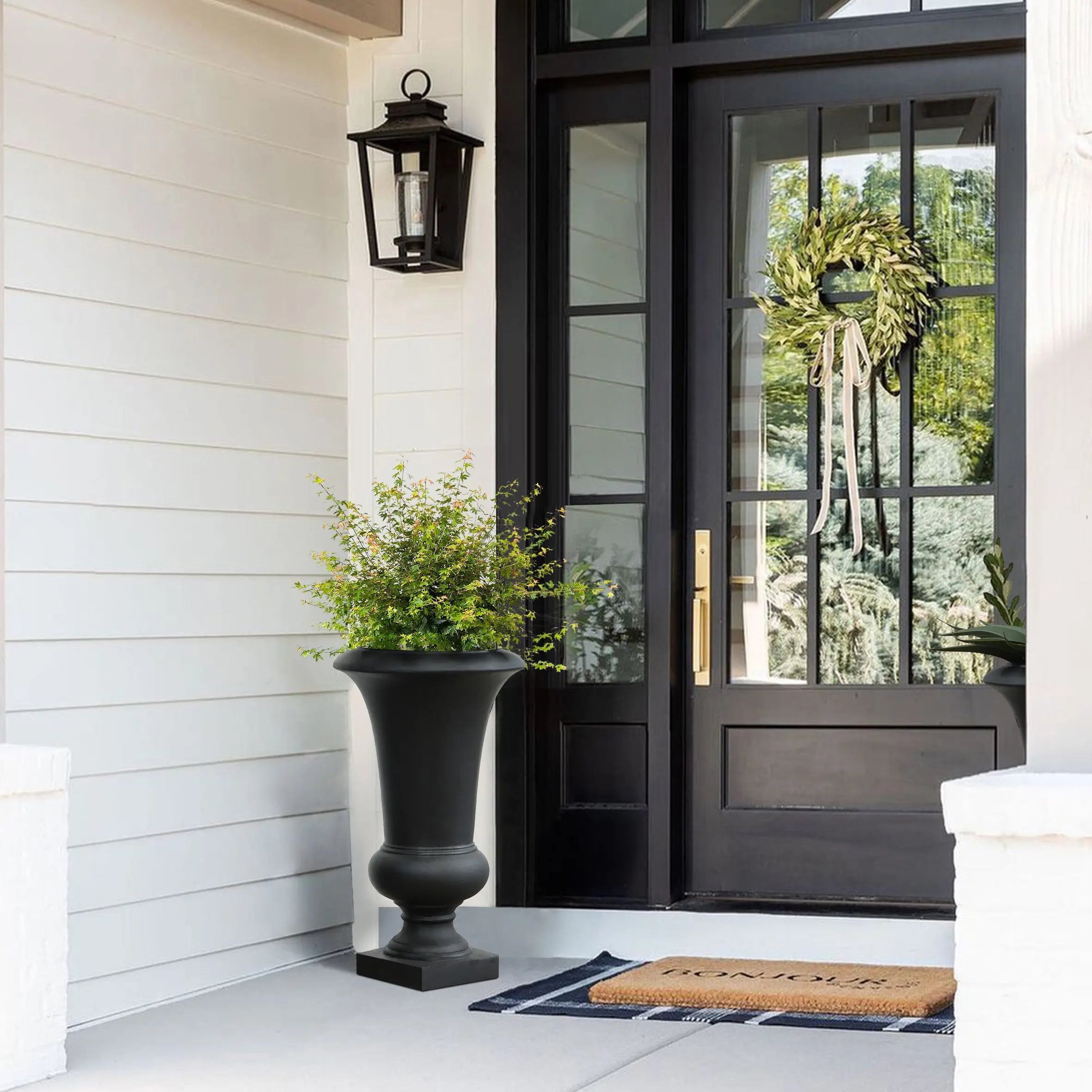 A weather-resistant black urn planter with green foliage sits next to a black-framed glass front door decorated with a wreath; a lantern is mounted above and a “welcome” doormat is on the porch.