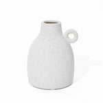 A vintage-inspired design emerges in this speckled white ceramic vase, featuring a rounded body and a loop handle on one side, set elegantly against a plain white background.