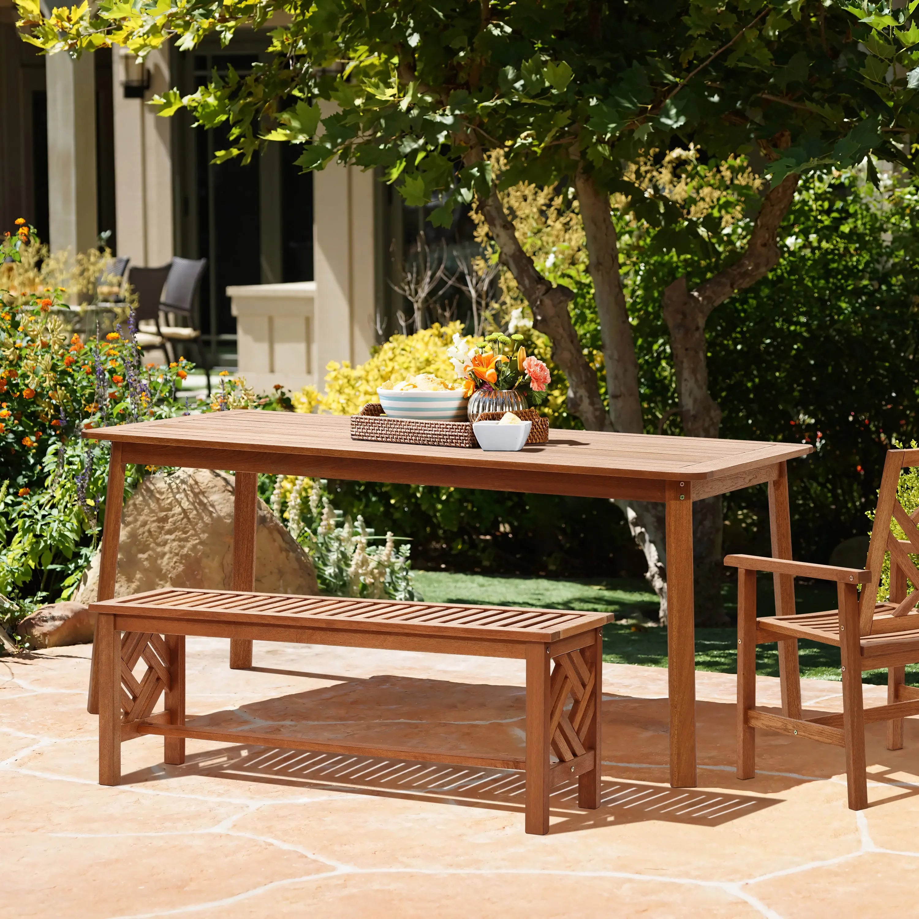 A solid Malaysian hardwood outdoor dining table set with a bench and chair graces the stone patio, surrounded by lush trees and plants. The table, treated with teak oil coating, features a vase of flowers and a tray, creating an inviting natural ambiance.
