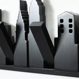 A metallic black sculpture depicting a stylized city skyline with various building shapes against a white background cleverly doubles as a wall-mounted coat rack, featuring discreet flip-down hooks to blend artistry and functionality seamlessly.