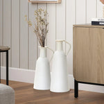 Two white ceramic vases with dried branches stand on a wooden floor next to a light wood cabinet in a room with vertical striped walls, embodying modern minimalist design. This harmonious home decor element is perfect for those who appreciate subtle elegance.