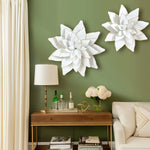 The room features green walls adorned with white lotus wall decorations, a wooden table, bottles, and a lamp. A bouquet of flowers graces the space alongside a white couch and an array of books, creating an inviting ambiance.