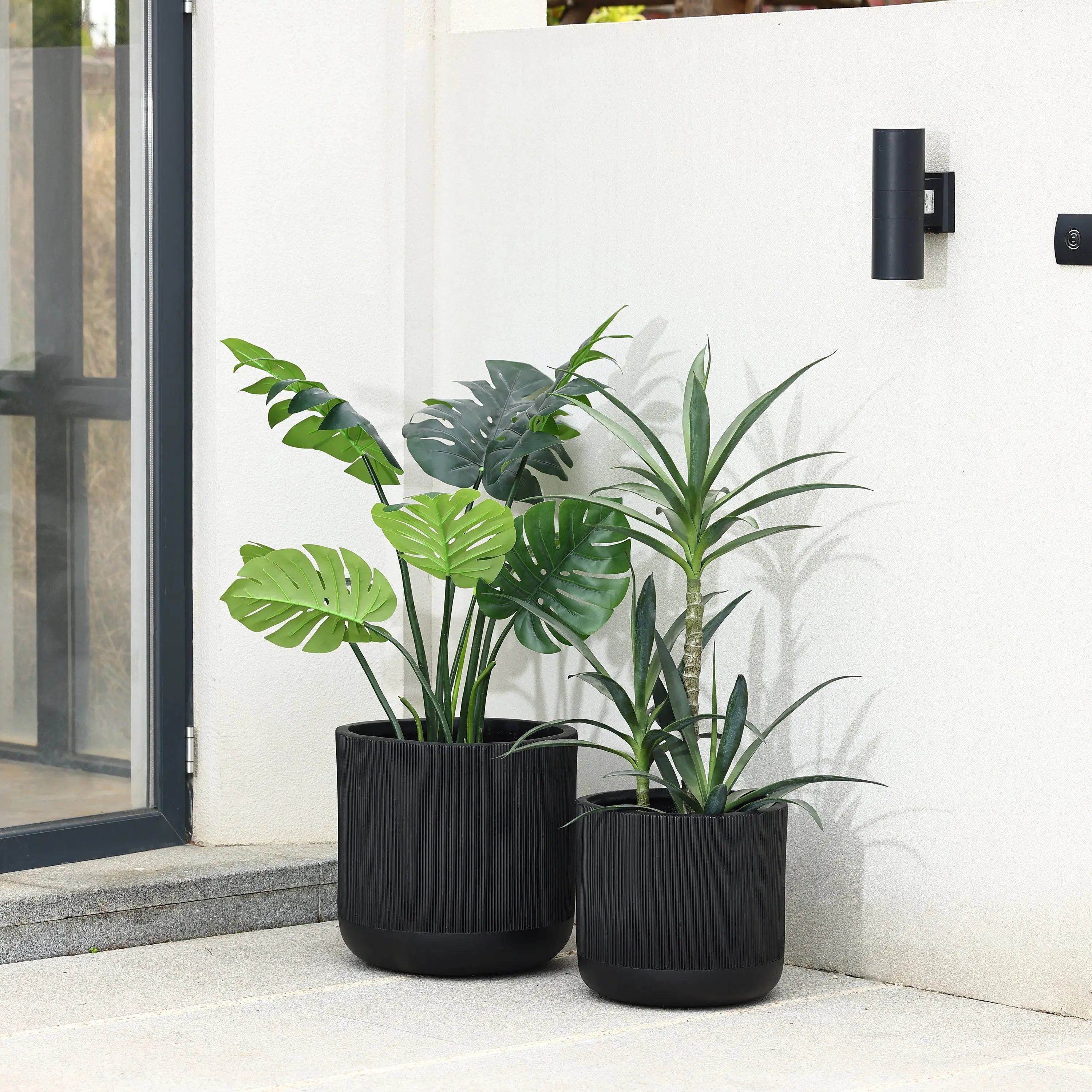 Two large black planters, made from durable magnesium oxide material, house green leafy plants. Theyre positioned against a white wall near a glass door and an outdoor light fixture.