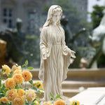 A white Mary statue, crafted from durable magnesium oxide, stands with outstretched hands near yellow roses in an outdoor garden setting.