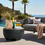 The outdoor seating area features a wicker sofa and a black cement outdoor coffee table, which holds a vase with flowers, a bottle, and two glasses. Palm trees sway gently against the backdrop of an ocean view.