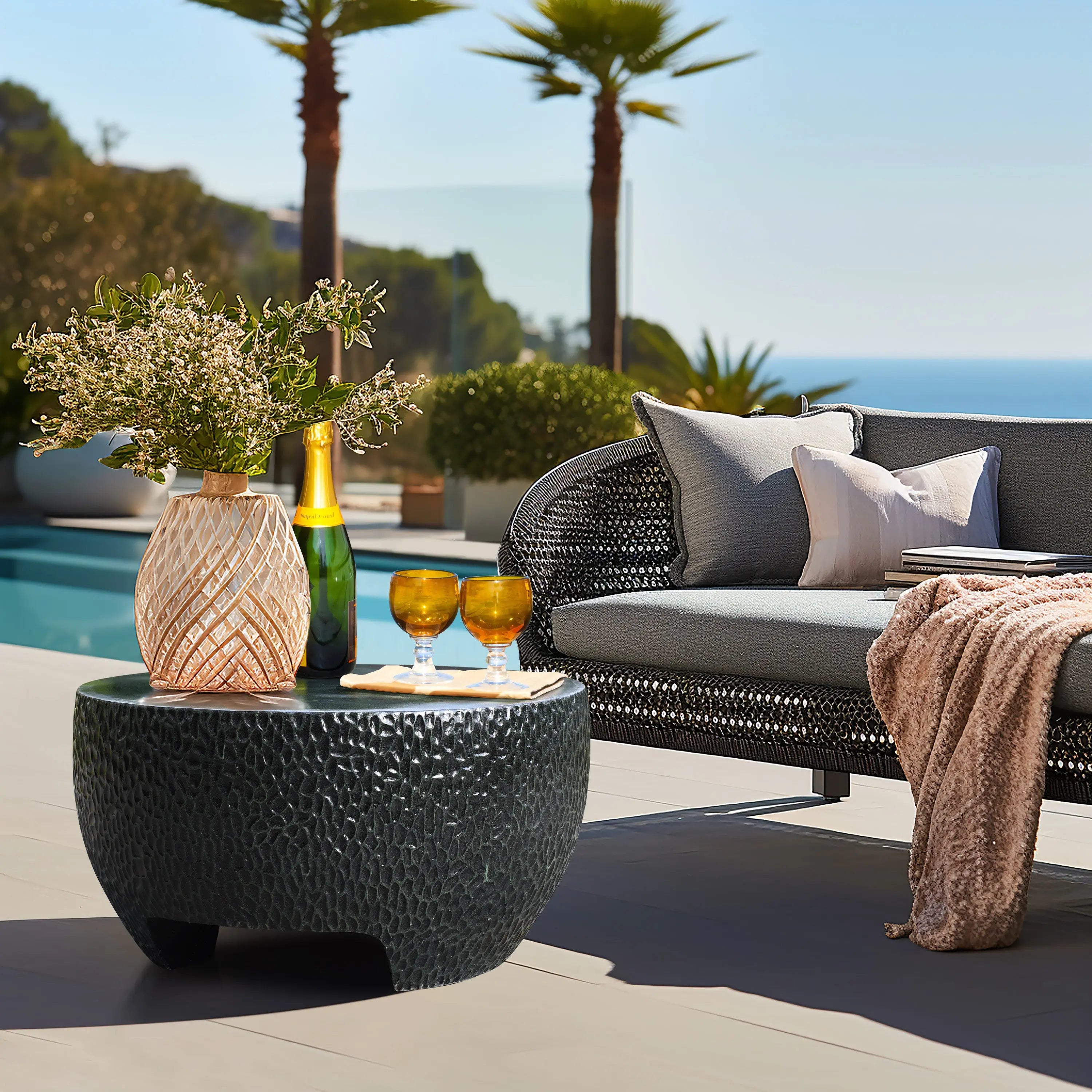 The outdoor seating area features a wicker sofa and a black cement outdoor coffee table, which holds a vase with flowers, a bottle, and two glasses. Palm trees sway gently against the backdrop of an ocean view.